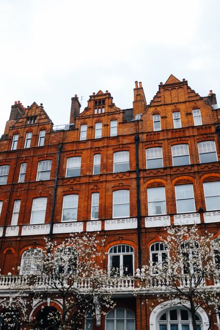 A multi-story red brick building with white-trimmed windows and ornate architectural details, featuring a white balcony on the second floor. The facade appears clean and well-maintained, with a few yo