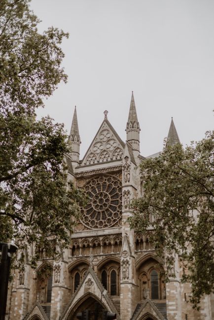 Photograph of the exterior of Westminster Abbey, showcasing its Gothic architectural style with tall, pointed spires, decorative stone carvings, and large stained glass windows. The building's surface